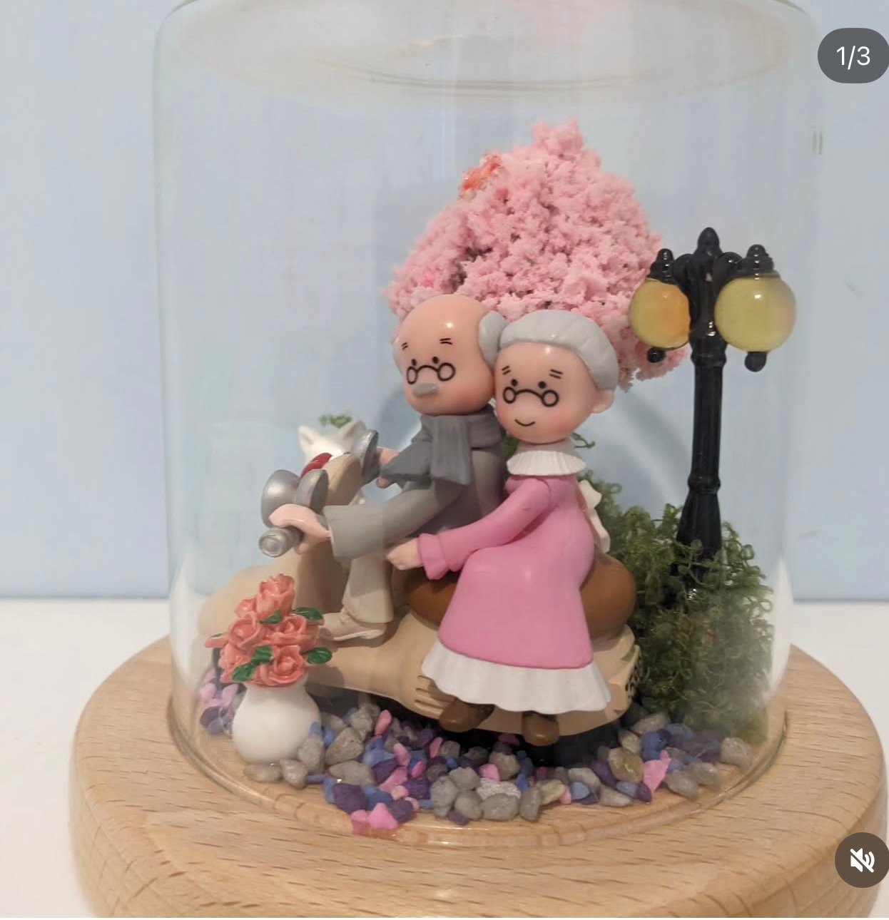 Couple de personnages sur un scooter sous une cloche, avec arbre rose et socle en bois